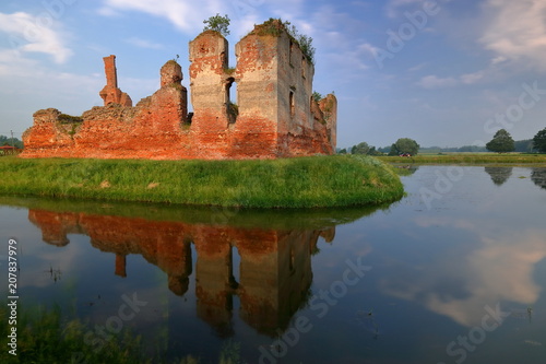 Malowniczy krajobraz z ruinami średniowiecznego zamku na porośniętej trawą wysepce otoczonej fosą z wodą, w której pięknie odbija się budowla oraz niebo, ciepłe światło wschodzącego słońca