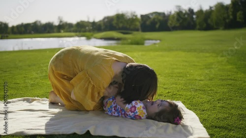 Funny mother and daughter having fun on green grass lawn. Young mom blowing into little girl's stomach tickling her. Toddler down syndrome daughter enjoying and laughing together with mother on meadow photo