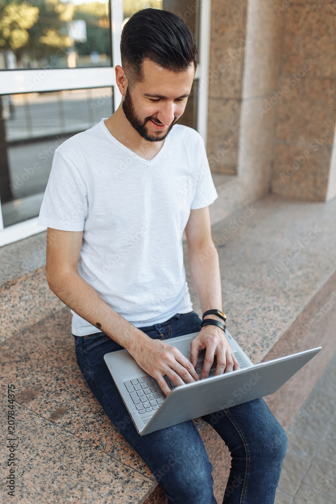 Portrait of a beautiful stylish guy dressed in a white t-shirt, who sits on a parapet, works with a laptop, on the street against a glass building, can be used for advertising, text insertion Portrait of a beautiful stylish guy dressed in a white t-shirt, who sits on a parapet, works with a laptop, on the street against a glass building, can be used for advertising, text insertion
