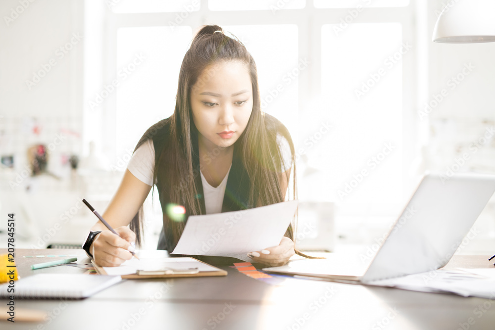 Portrait of focused creative Asian woman drawing sketches for while standing at work table in sunlit fashion design atelier, lens flare Portrait of focused creative Asian woman drawing sketches for while standing at work table in sunlit fashion design atelier, lens flare