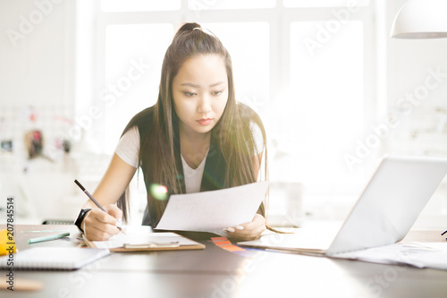Portrait of focused creative Asian woman drawing sketches for while standing at work table in sunlit fashion design atelier, lens flare