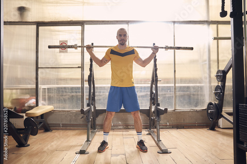 one young man, wearing sport clothes, about to do squat exercise with bar, in old beaten up gym interior. full lenght shot.