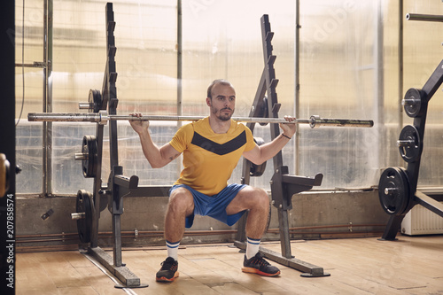 one young man, wearing sport clothes, squat exercise with bar, in old beaten up gym interior. full lenght shot. photo