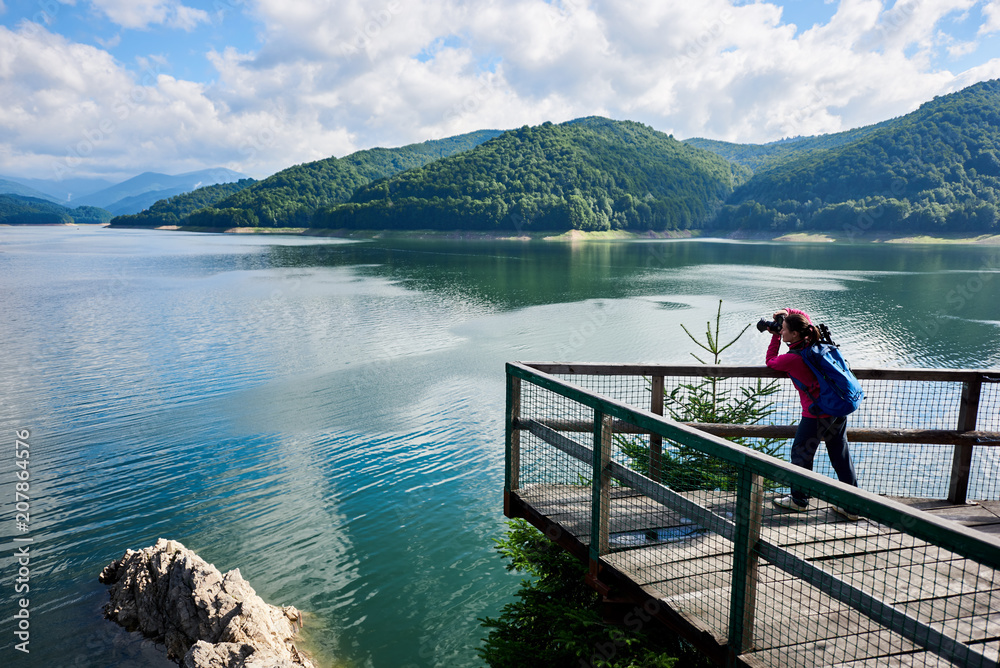 Young female photographer with a blue backpack on the lake. Beautiful scenery of a sunny day: mountains, forests, lake, blue sky with clouds