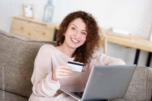 Happy and joyful redhead woman looking at camera with happy expression  hold in hand credit card  making transaction using laptop pc. Online shopping. People  business and finances concept.