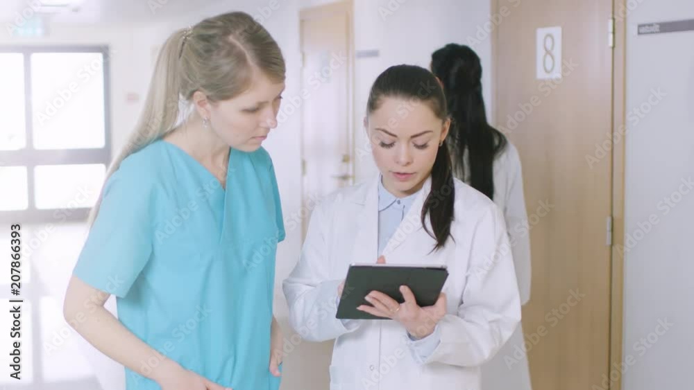 In the Hospital, Busy Doctors Talk, Using Tablet Computer While Standing in the Hallway. New Modern Fully Functional Medical Facility. Shot on RED EPIC-W 8K Helium Cinema Camera. In the Hospital, Busy Doctors Talk, Using Tablet Computer While Standing in the Hallway. New Modern Fully Functional Medical Facility. Shot on RED EPIC-W 8K Helium Cinema Camera.
