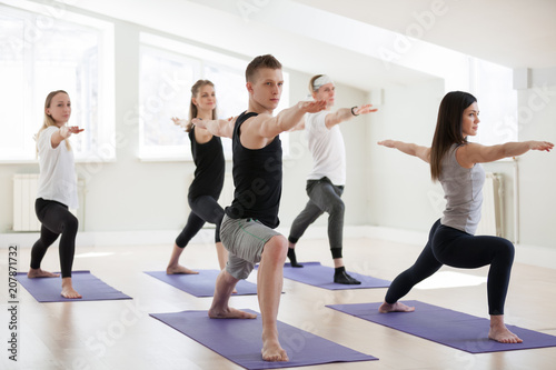 Group of young sporty people practicing yoga lesson, doing Warrior Two exercise, Virabhadrasana 2 pose, working out, indoor session full length, students training in club, studio. Wellbeing lifestyle
