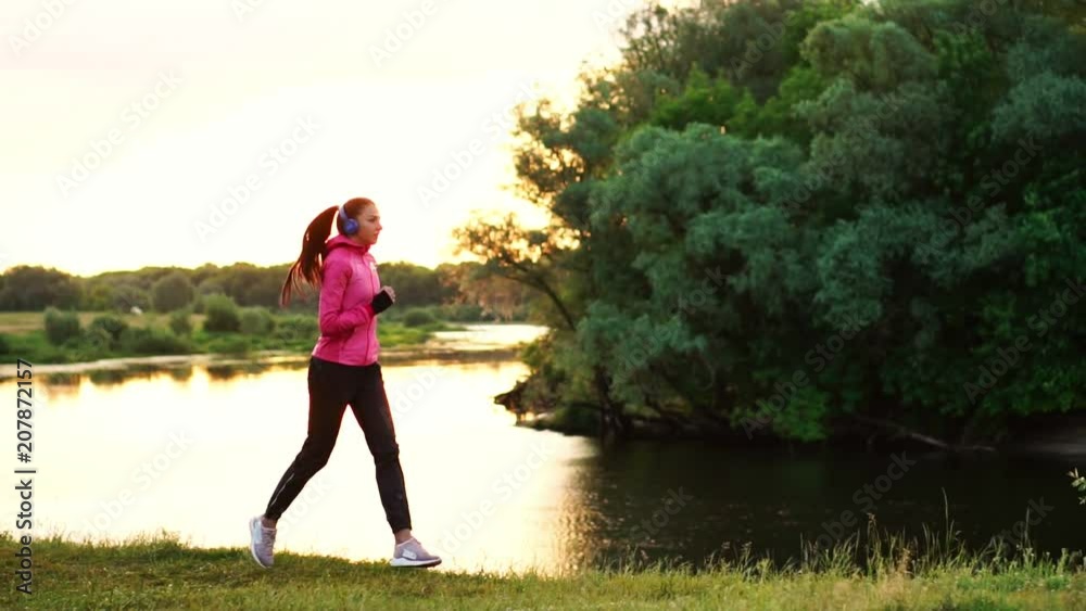 A girl in a pink jacket and black pants runs near the river in headphones preparing for the marathon A girl in a pink jacket and black pants runs near the river in headphones preparing for the marathon