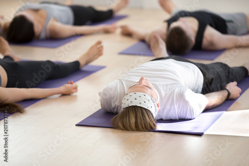 Group of young sporty people practicing yoga lesson, doing Savasana pose, Corpse, Dead Body exercise, working out, indoor full length, students training in sport club