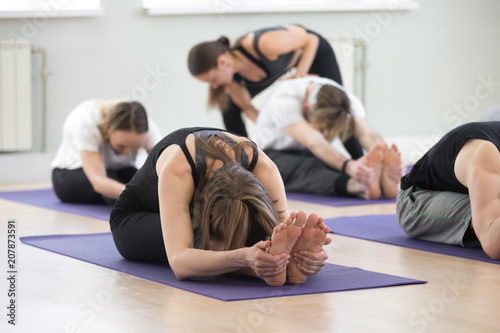 Young female yoga instructor teaching paschimottanasana pose, Seated forward bend exercise for a group of sporty people practicing in studio, working out indoor, teacher helping to master, full length