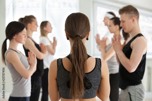 Group of young sporty people practicing yoga with instructor, relax after lesson or starting class, doing meditation exercise, Namaste pose, indoor close up, yogi students working out in sport club