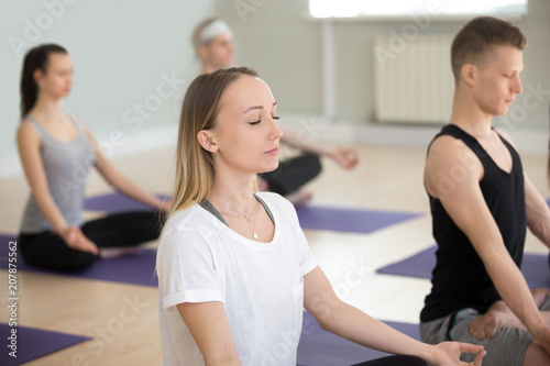 Group of young sporty people practicing yoga lesson, sitting in Sukhasana exercise, doing Easy Seat pose, working out, indoor close up, students having sport class, training in club, studio