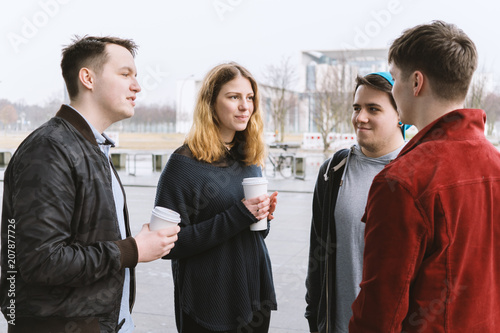 group of teenage friends having a conversation while standing together on city street holding coffee cups