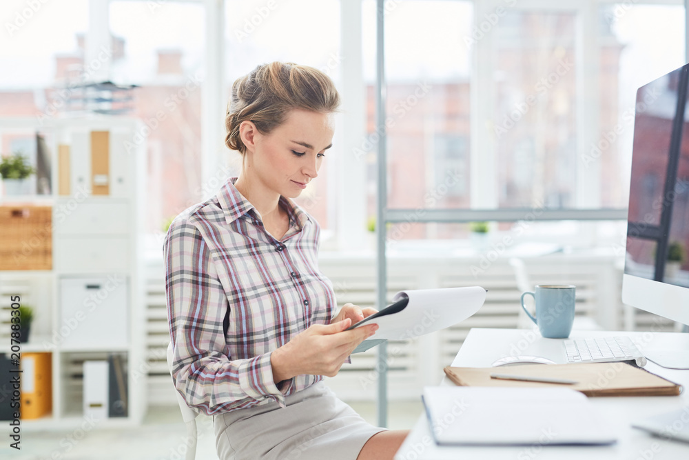 Obraz premium Young pretty Caucasian woman sitting at office desk and reading important paper on clipboard