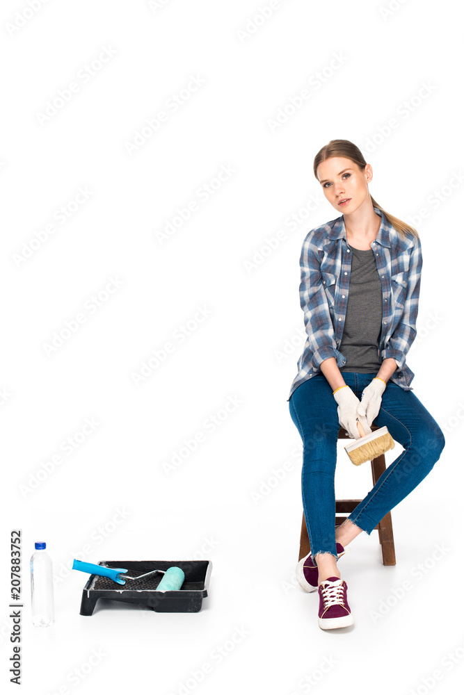 young woman sitting on chair with paint brush near bottle, roller tray and paint roller isolated on white background young woman sitting on chair with paint brush near bottle, roller tray and paint roller isolated on white background