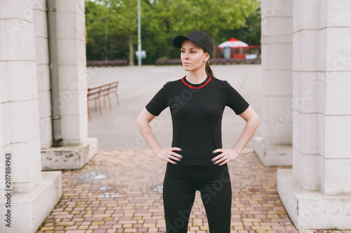 Young concentrated athletic beautiful brunette girl in black uniform and cap doing sport exercises, warm-up before running, standing in city park outdoors. Fitness, healthy lifestyle concept. © ViDi Studio