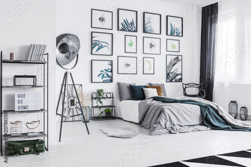 Real photo of a cozy bed standing next to a black lamp in a monochromatic bedroom with shelves with ornaments and posters on a wall