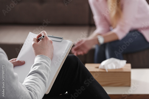 Photography Close-up of therapist hand writing notes during a counseling session with a single woman sitting on a couch in the blurred background