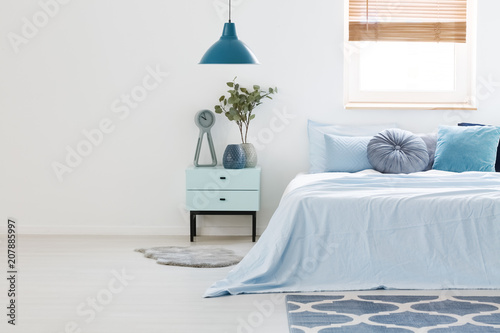 Real photo of a blue and white bedroom interior with wooden nightstand between a light blue bed and an empty wall. Paste your armchair here