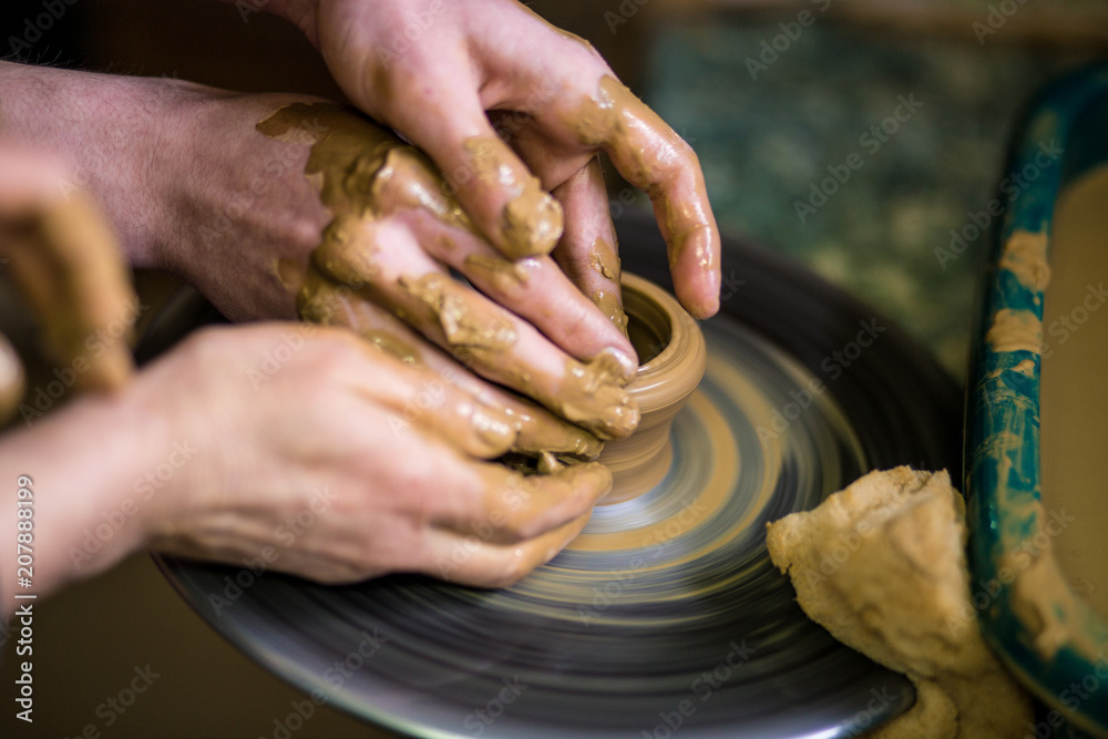 Close-up hands of potter in apron making vase from clay, selective focus. Making it together. Top view of potter teaching to make ceramic pot on pottery wheel