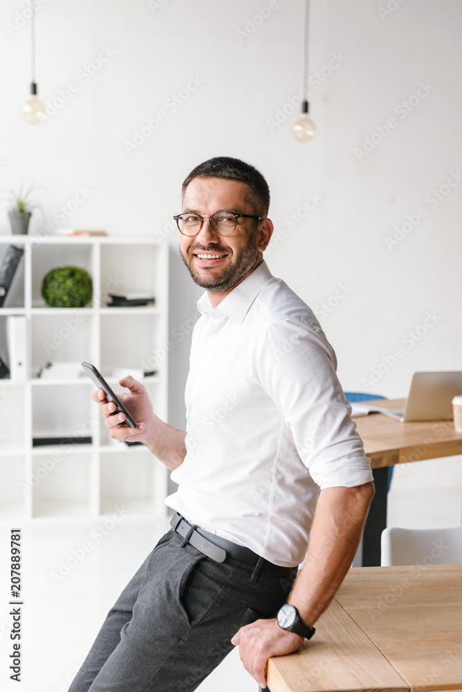 Portrait of attractive european man in formal wear sitting on table in office and looking at you, while holding smartphone Portrait of attractive european man in formal wear sitting on table in office and looking at you, while holding smartphone