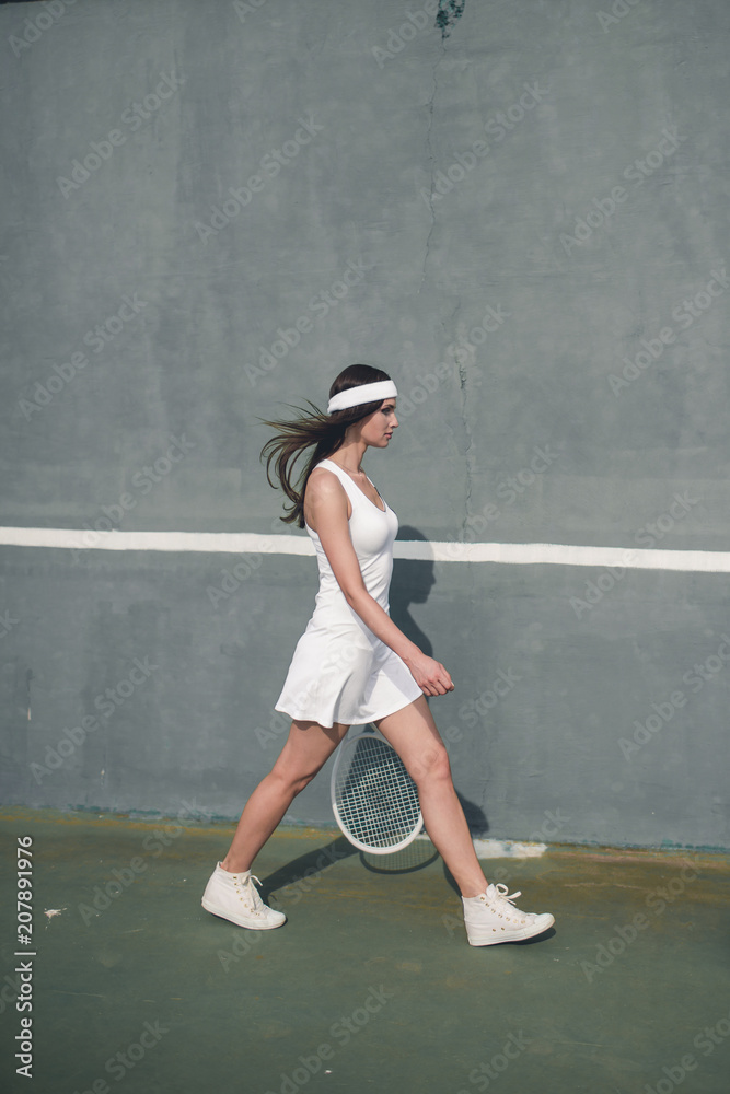 Portrait of young Caucasian teen model wearing fashionable tennis dress, walking on tennis hardcourt, summer sunny day outdoors. Fashion portrait shoot