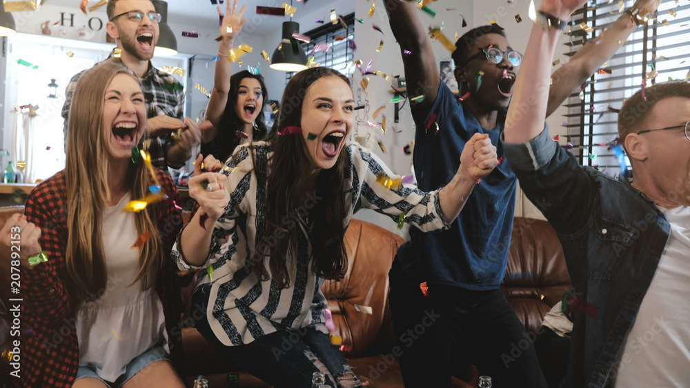 © vadim_key - Emotion. Multi-ethnic fans celebrate winning. Confetti 4K slow motion. Passionate supporters shout watching game on TV.