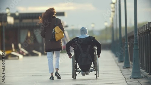 Young happy woman with disabled man in a wheelchair walking together on the quay