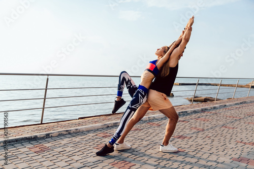 Healthy lifestyle. Young woman and man doing sports exercises for body together, wearing sportswear, while working out near the sea, on the quay.