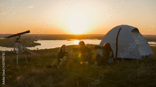 Group of friends sitting on the grass near the tent chilling out at sunset