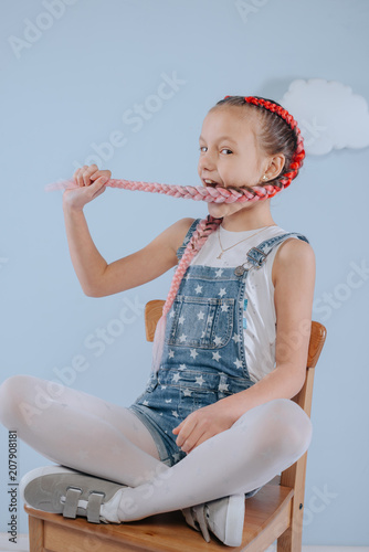 Stylish teenage girl is enjoying her modern kanekalon hair. little girl in fashionable denim overalls.girl sits on a wooden chair in the studio