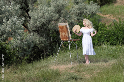 Beautiful girl draws a picture in the park using a palette with paints. Easel and canvas with a picture. photo