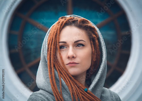 Portrait of a girl with dreadlocks of red color in a white sweatshirt with a hood on the background of a large round window photo