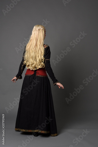 full length portrait of pretty blonde lady wearing  a red and black fantasy medieval gown. standing pose on grey background.