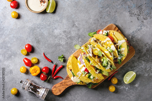 Variety of vegetarian corn tacos with vegetables, green salad, chili pepper served on olive wood board with tomato, cream sauces with ingredients above over grey texture background. Top view, space.