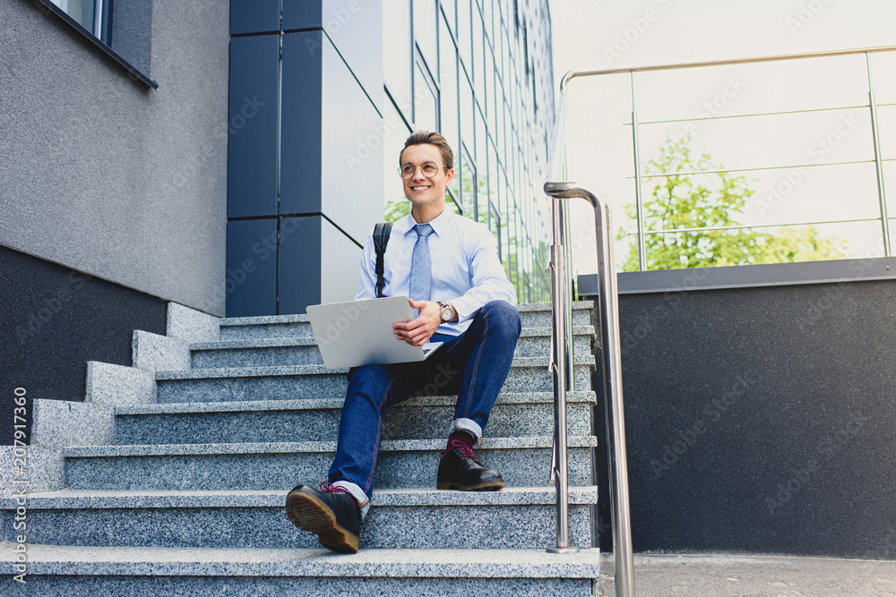 handsome smiling young freelancer in eyeglasses sitting on stairs and using laptop