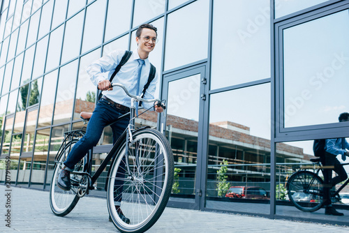 handsome smiling young man in eyeglasses and formal wear riding bike on street