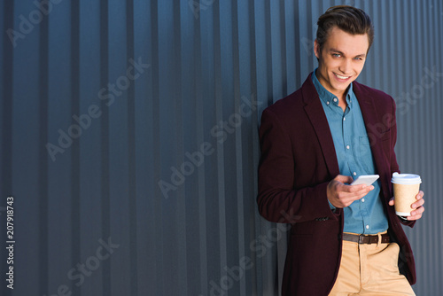 handsome young man with coffee to go using smartphone and smiling at camera