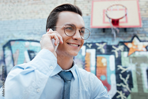 smiling young businessman in eyeglasses talking by smartphne and looking away on street