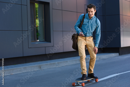 smiling young man riding longboard and using smartphone