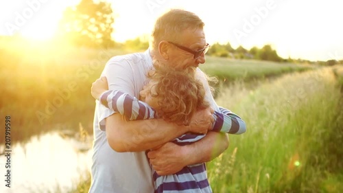 Elderly grandfather and loving grandson smiling and hugging each other outdoors, on the river bank. Two generations. Colorful sunset. A happy family