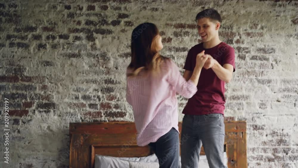 Happy lovers are dancing on bed holding hands and enjoying married life together. Modern double bed, brick wall and light loft apartment interior is visible. Happy lovers are dancing on bed holding hands and enjoying married life together. Modern double bed, brick wall and light loft apartment interior is visible.