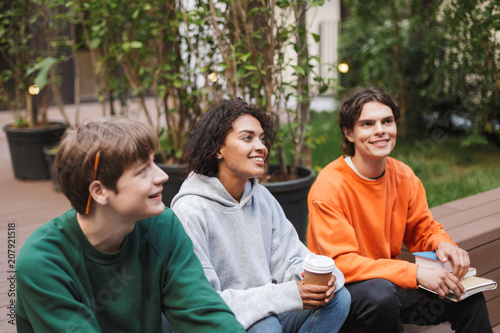 Group of smiling students sitting with coffee to go and books in hands and dreamily looking aside while studying together in courtyard of university