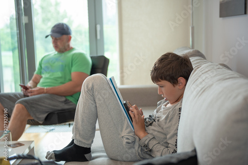 Young boy sitting in living room, looking to smartphone and drawing, with his father in the background 
