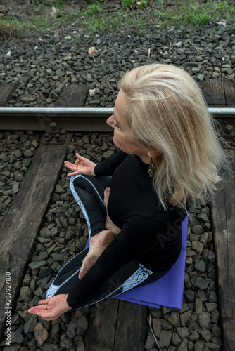 Attractive senior blond woman, dressed in black, doing yoga poses outdoor on railroad. Concept: on the right path. 