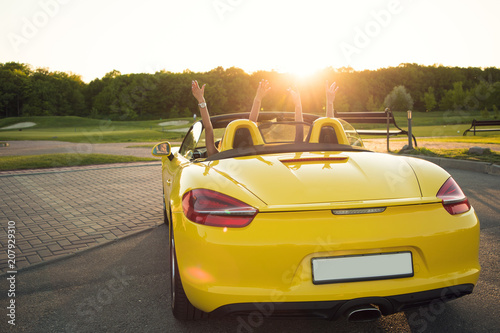 Follow the luxury way of life  Back view photo of two young wealthy stylish girls raising their hands upward  while sitting in the cabrio in the sun shine.