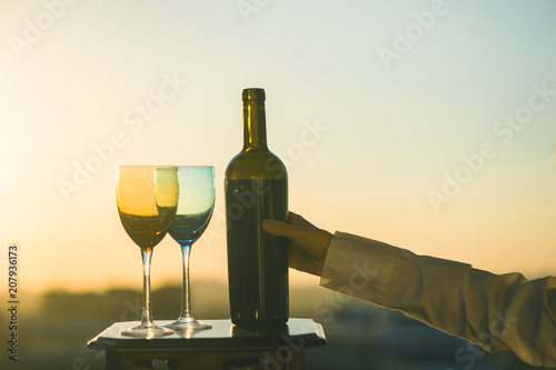 Female hand with bottle pours red wine into glasses on blurred night city background. Service on the roof of the restaurant