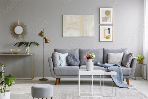 Real photo of a great sofa with cushions and blanket standing in elegant living room interior behind a white table and next to a gold lamp and grey wall with posters