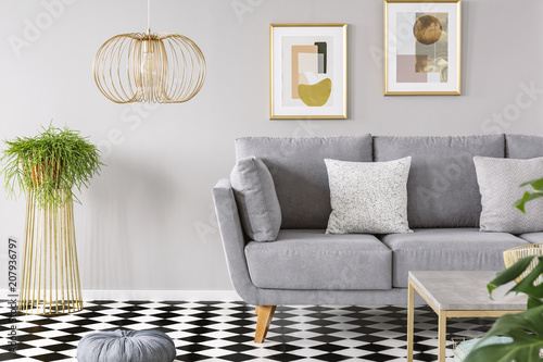 Real photo of a living room interior with gold posters on the walls, grey sofa with cushions and checkered floor