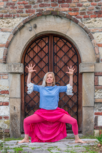 Goddess Yoga Pose – Deviasana. Attractive blond mature woman doing yoga poses outdoor,  in front of old fortress. Longevity concept